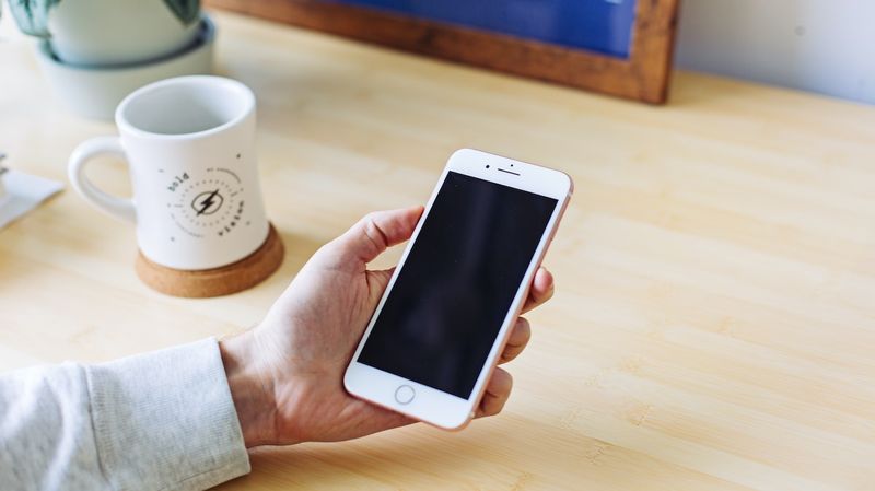 A hand holding an iPhone over a wooden desk, with a cup of coffee to the side and a framed photo in the background