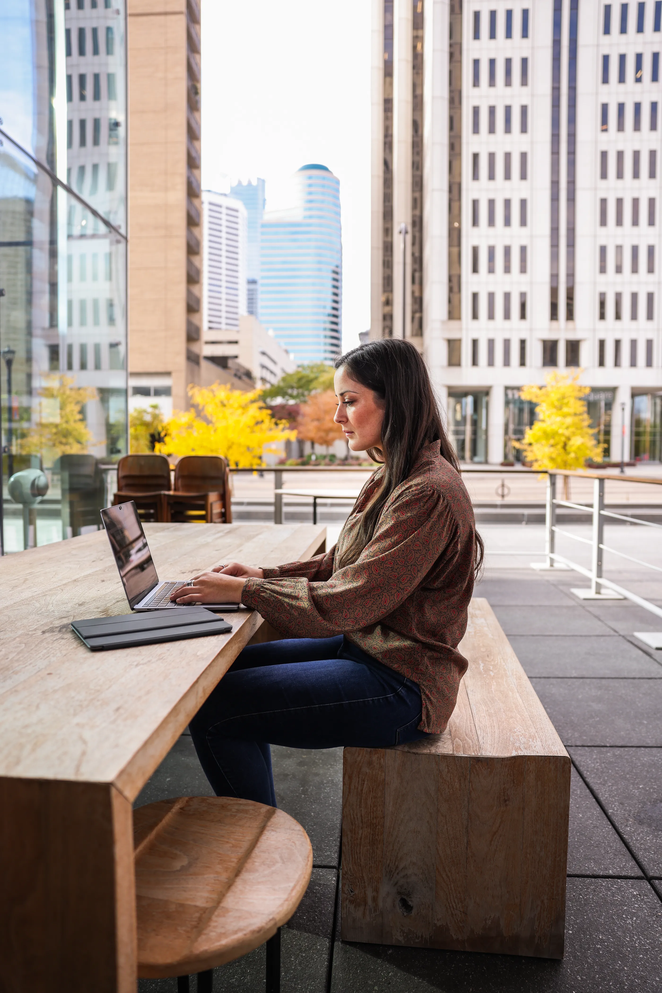 A woman with long dark hair is working on a laptop at a wooden table outdoors in a city setting.