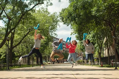 Four people jump in the air on a park path, holding blue foil balloons that spell out