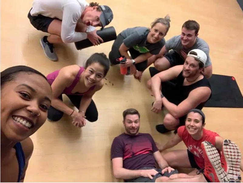 A multi-ethnic group of Jamfs look up, smiling, from a gym floor. They are wearing workout clothes.