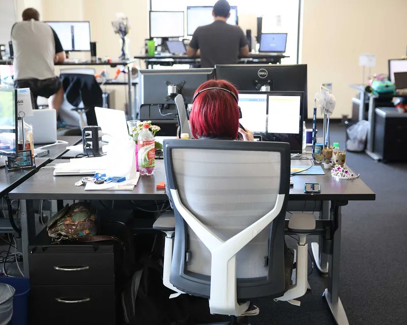 A person with short, bright red hair is sitting in an office chair at a desk with multiple computer monitors, working in a busy office environment.
