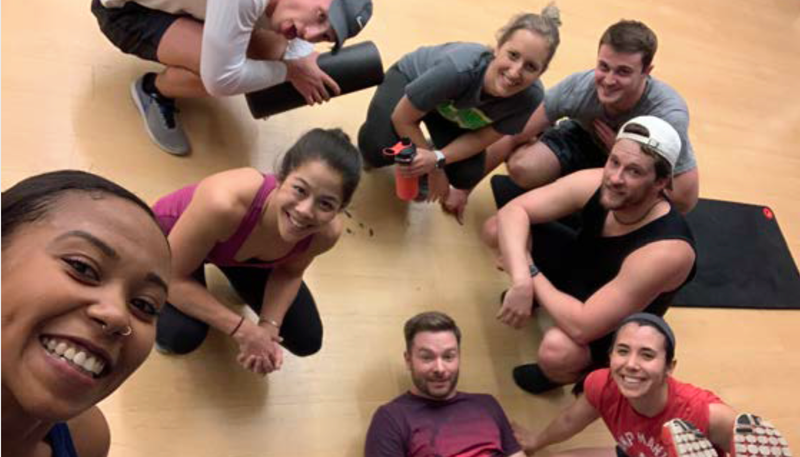 A diverse group of seven smiling people are gathered in a circle on a wooden floor, possibly after a workout.