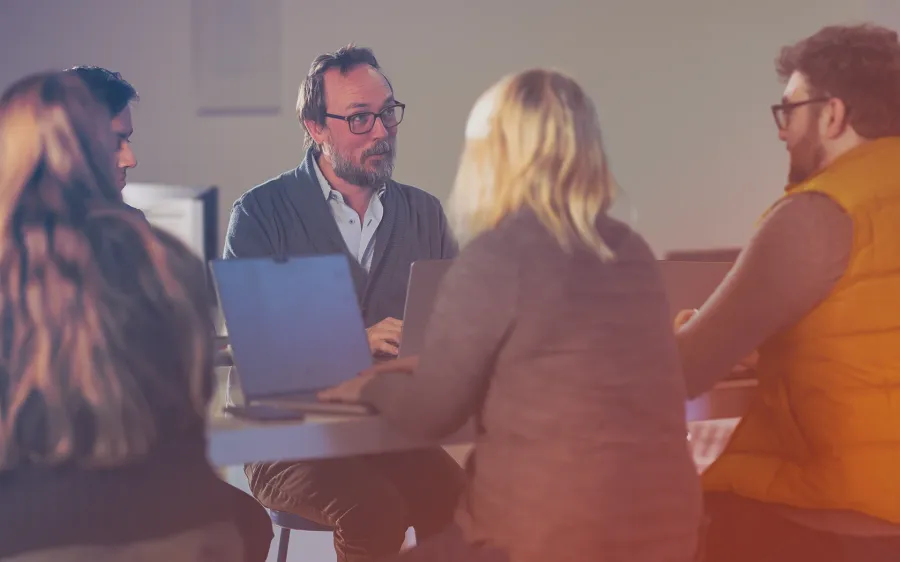 A male Jamf employee with glasses and a beard speaks to customers during a collaborative session in an office. They are seated at a table with open Apple Macbooks, engaged in conversation.