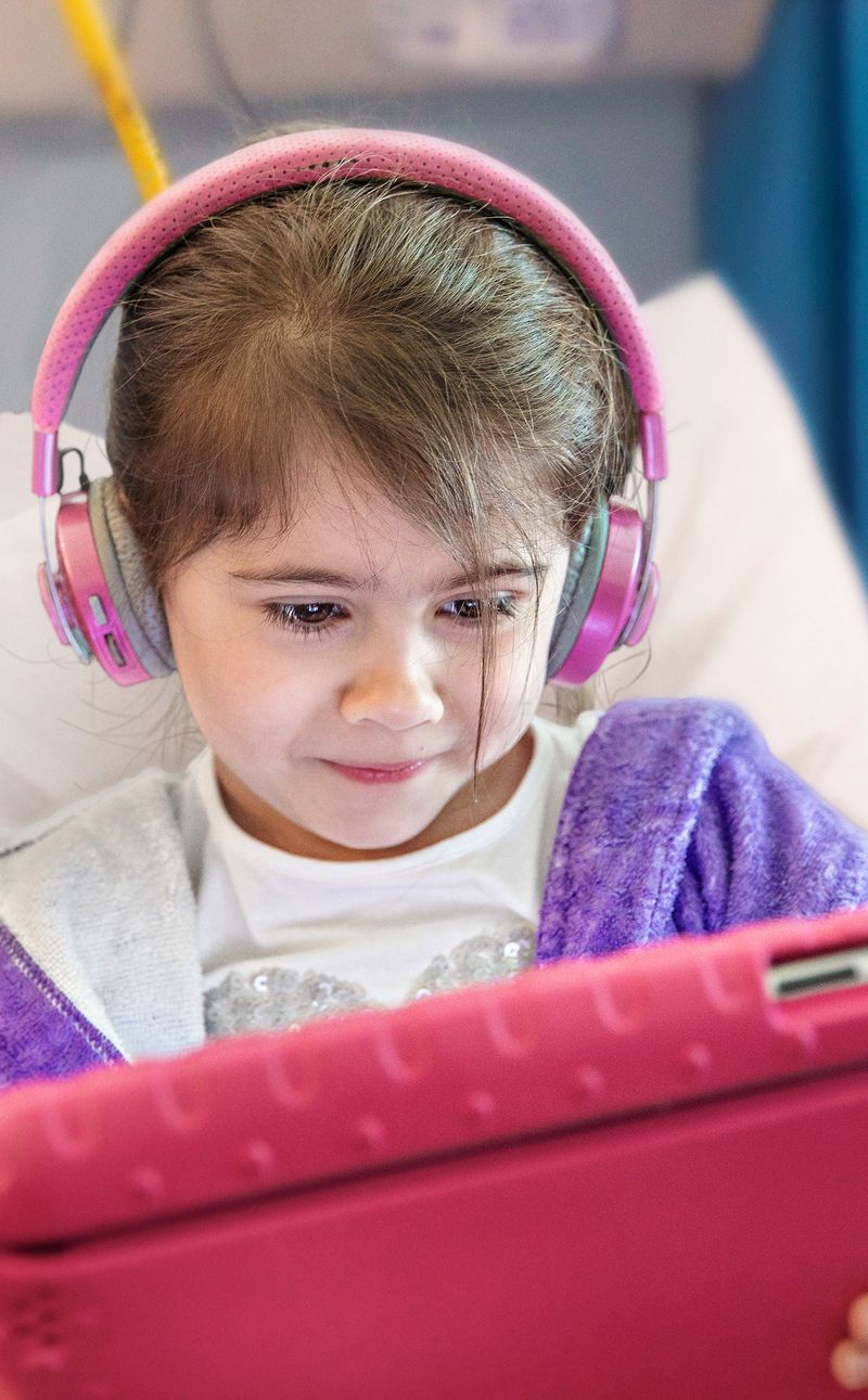 Child in a hospital bed wearing headphones and using an iPad.