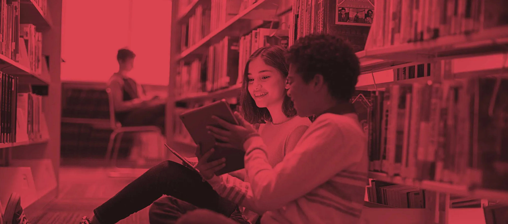 Two kids, a boy and a girl, sitting on the floor of a library. The boy is showing the girl something on an iPad.