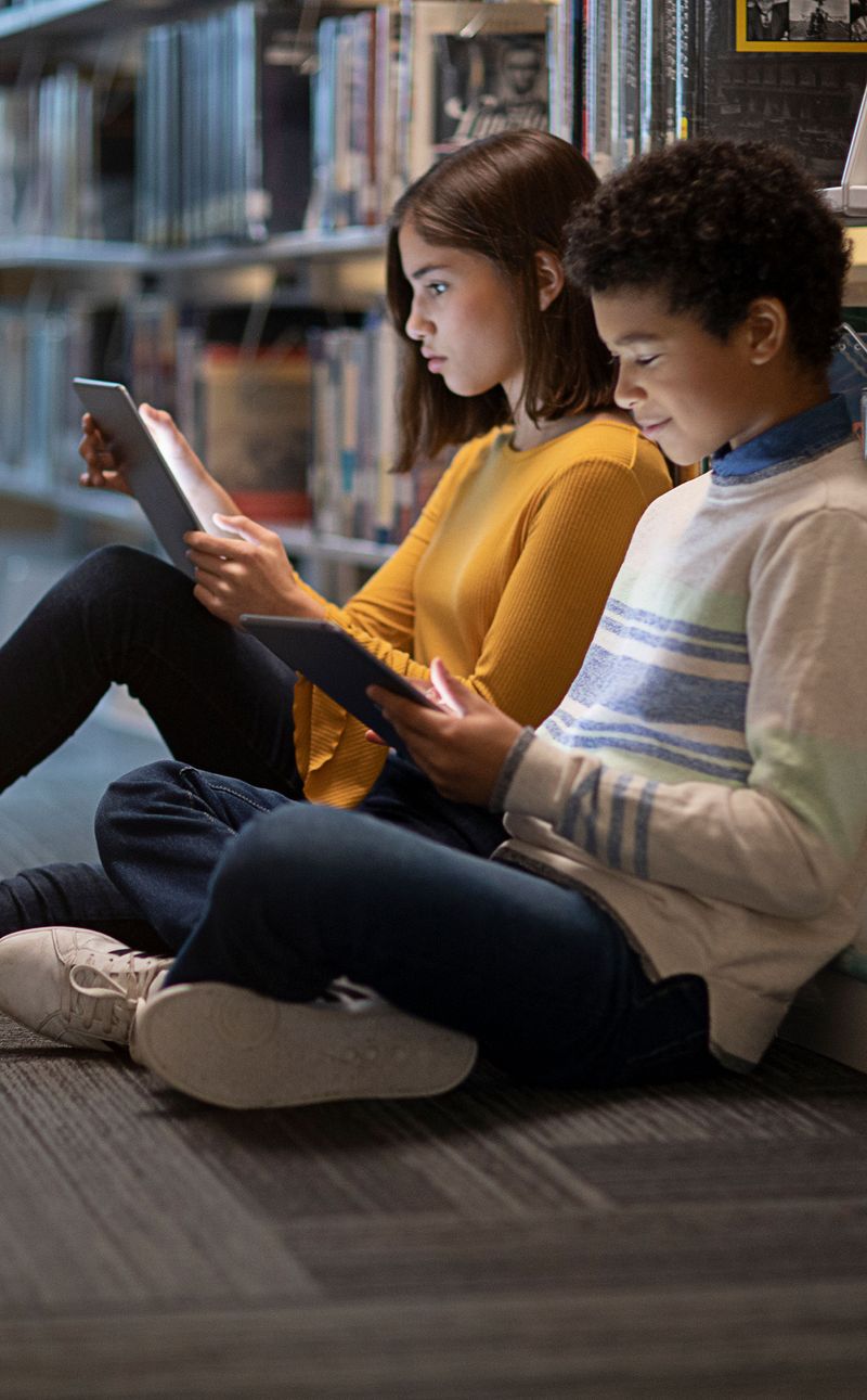 Two children sitting on the floor of a library, leaning against the bookshelves. Both are looking at iPads.