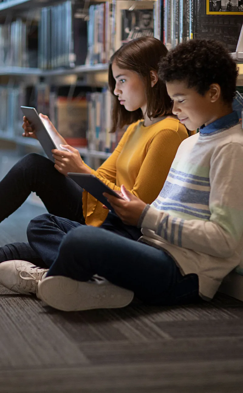 Two children sitting on the floor of a library, leaning against the bookshelves. Both are looking at iPads.