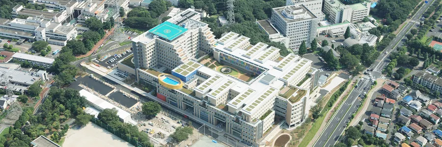 Aerial view of a large modern Japanese hospital campus with helipad, green rooftops, and surrounding urban infrastructure.