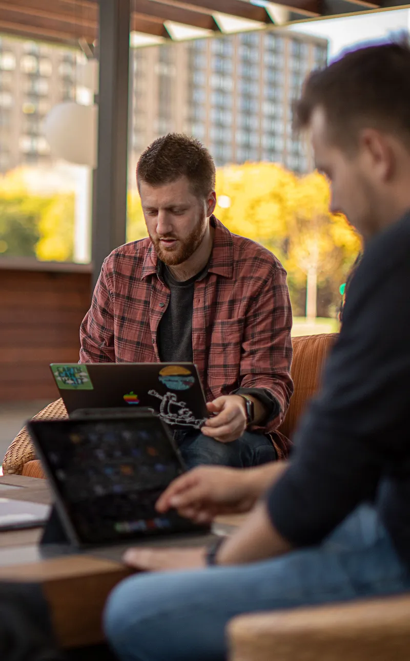 Two college students sitting outside on a patio. One has a MacBook in his lap, while the other is working on an iPad.