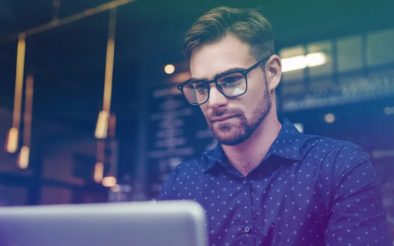 A focused man with glasses works on an Apple Macbook in a cafe with warm lighting.