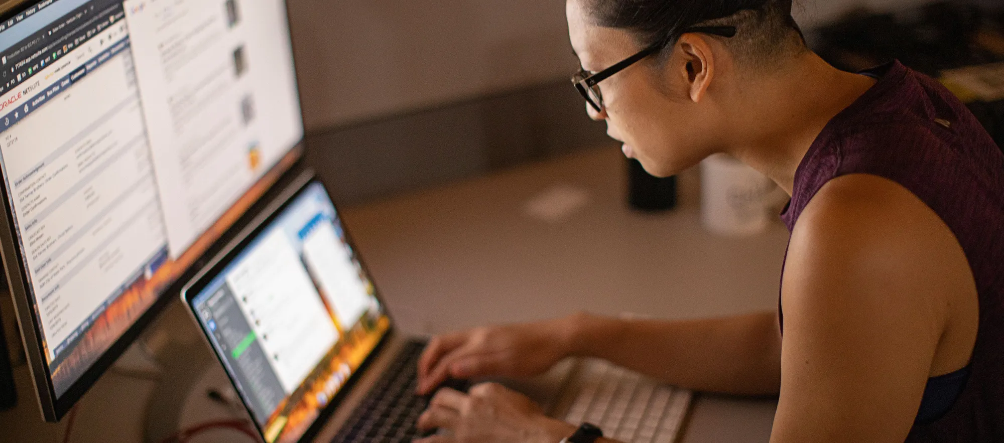 Person sitting at a desk working on a MacBook with a larger monitor behind the MacBook