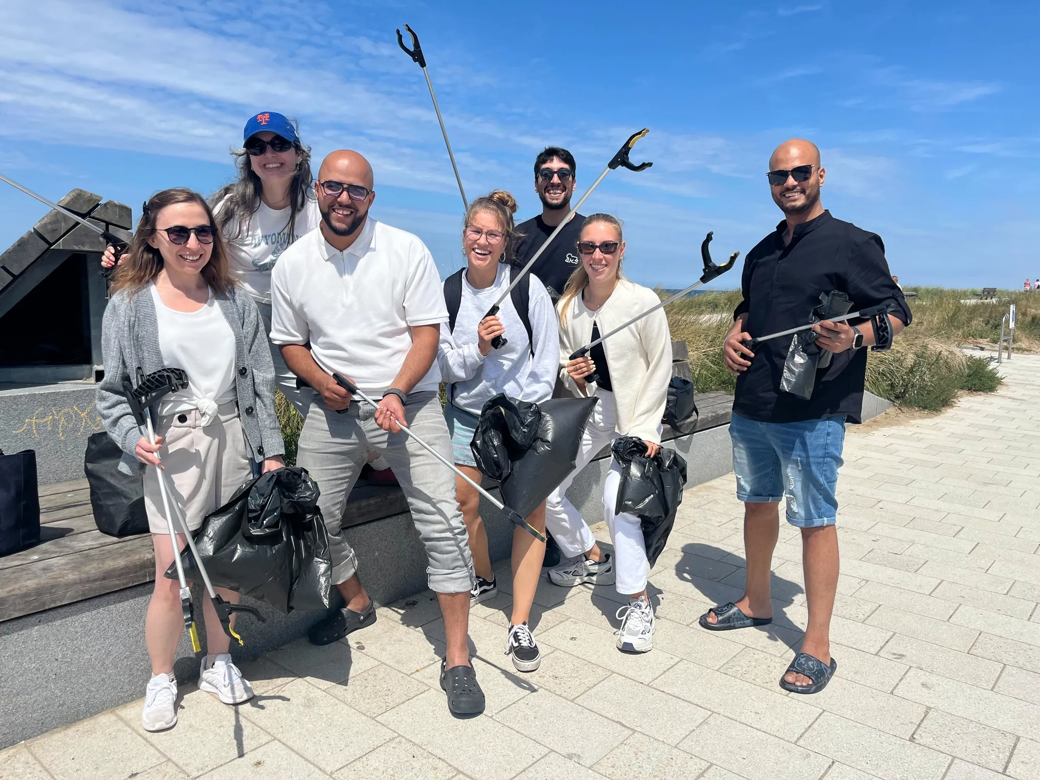 A group of seven Jamf volunteers holding trash grabbers and black plastic bags on a sunny beach boardwalk.