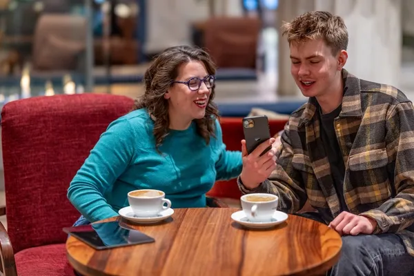 Two colleagues at Jamf sit at a round wooden table, smiling and looking at a smartphone together over cups of coffee.