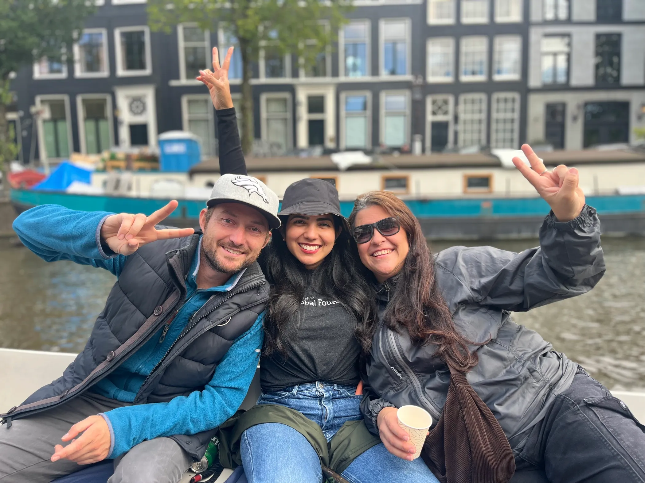 Three smiling Jamf colleagues sitting together on a boat in an Amsterdam canal, making peace signs with the city's historic architecture and a houseboat in the background.