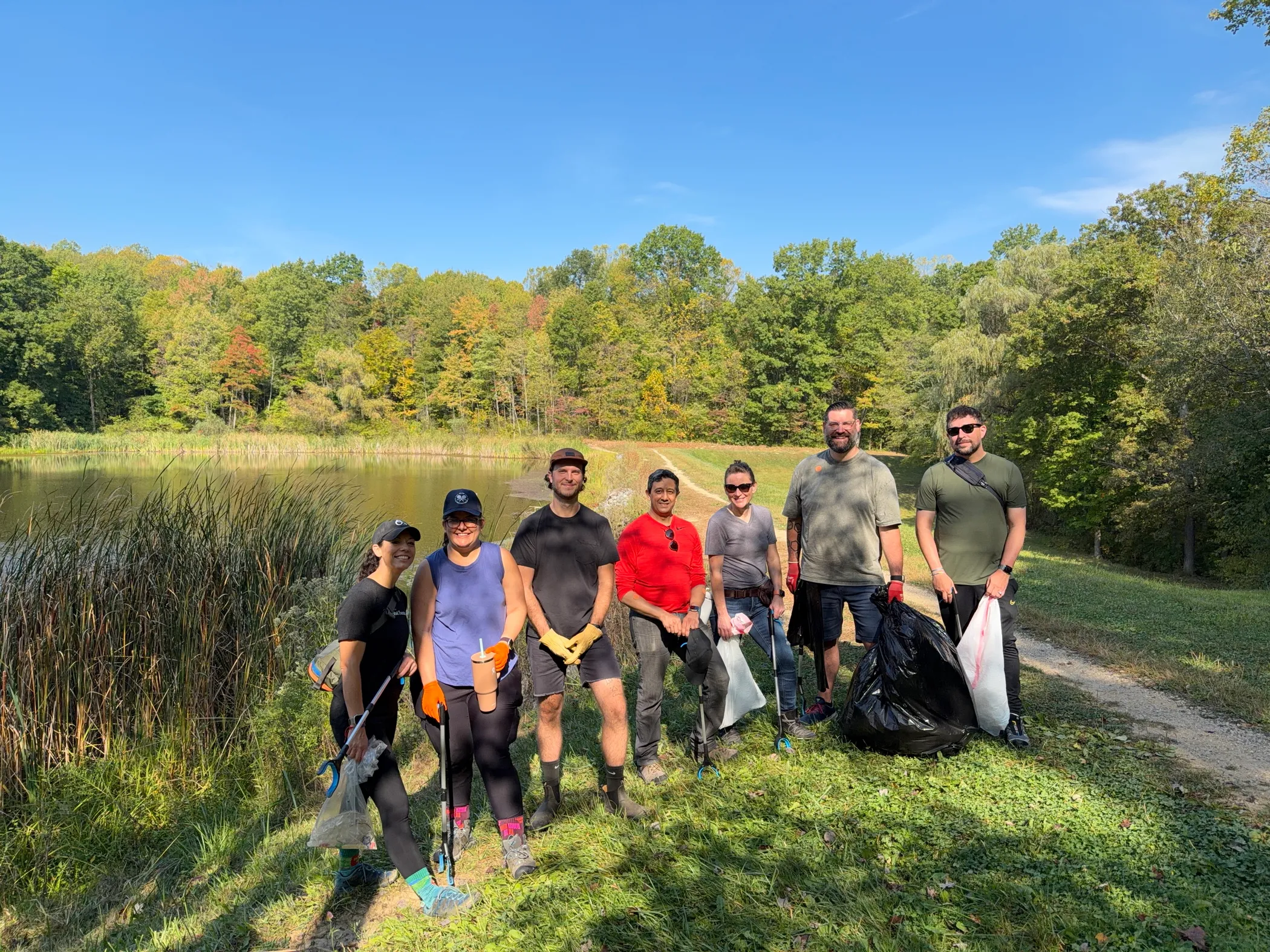 A group of seven Jamf volunteers standing on a grassy path by a lake and dense forest, holding trash grabbers and bags for a World Cleanup Day event in an Ohio national park.