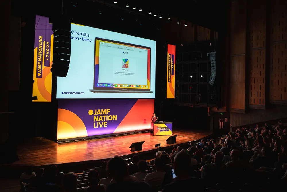 A speaker presents on a large stage at Jamf Nation Live London to a full auditorium of attendees.