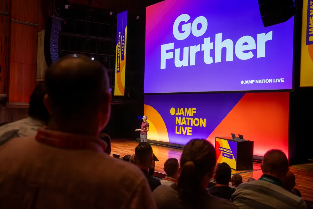 A speaker presents on stage at the Jamf Nation Live London event in front of an audience, with a large screen displaying the text