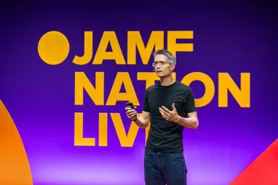 A speaker on stage at a Jamf Nation Live event. A man in a black t-shirt and glasses gestures while presenting in front of a vibrant purple and orange background featuring the 'JAMF NATION LIVE' logo in bold yellow typography.