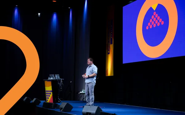A Jamf employee delivers a keynote at the Jamf Nation Live event from a dark stage with blue lighting and an overhead screen with a graphic.
