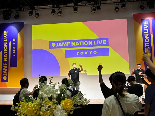 Jamf Nation Live Tokyo opening toast on stage with speaker and attendees raising drinks before a colorful branded backdrop