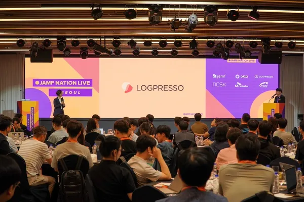 A wide shot of the audience seated at a Jamf Nation Live Seoul event. A speaker stands at a podium on the left side of the stage, and a large screen shows the Logpresso logo, with several sponsor logos to the right, including Jamf, Okta, and Logpresso.