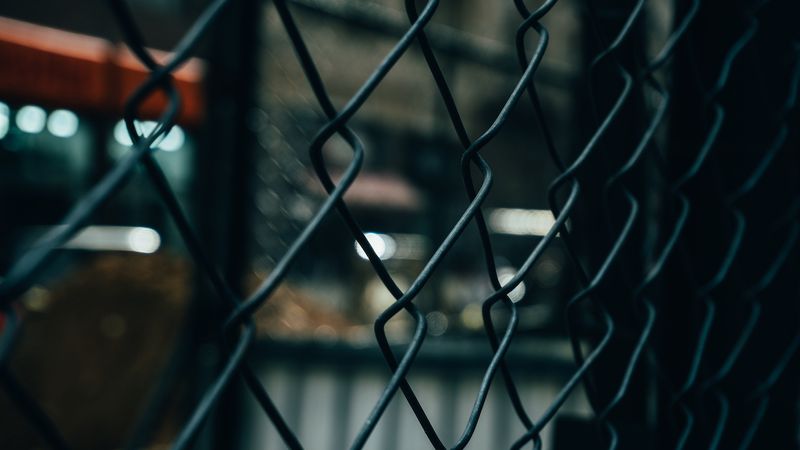 A chain-link fence with windows visible but out of focus behind it