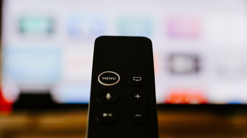 A black Apple TV remote in the foreground, with an out-of-focus display behind it