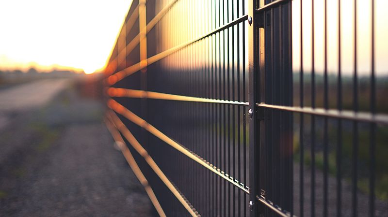 A close-up shot of a metal fence, running along a strip of gravel between a road on the left and a field on the right, under a setting sun. The fence symbolizes security with Jamf and Cisco.