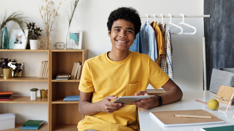 A teenage boy sitting at a desk inside a home, holding an iPad and smiling at the camera