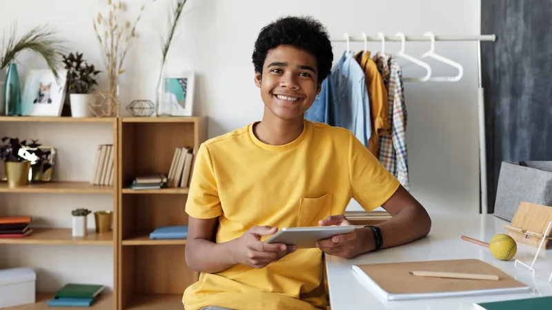 A teenage boy sitting at a desk inside a home, holding an iPad and smiling at the camera