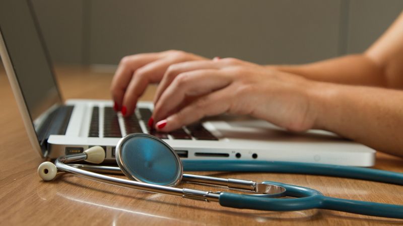 Hands typing on a MacBook with a stethoscope sitting adjacent