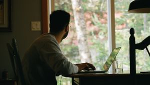 A man sitting at a table using a laptop while looking out the window at trees outside