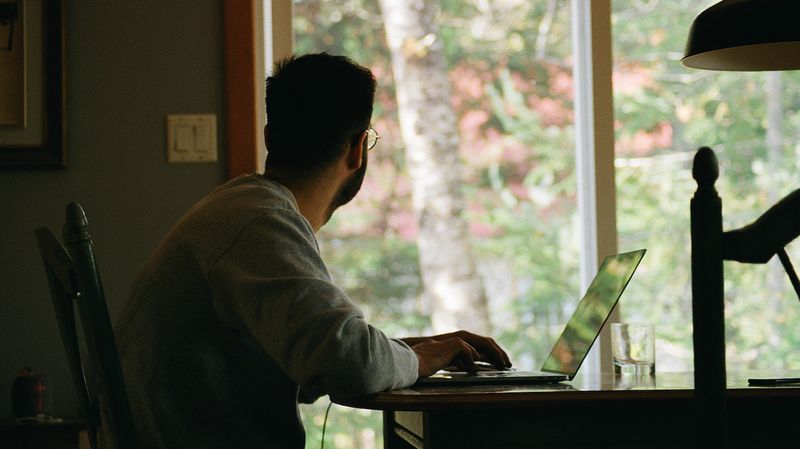 A man sitting at a table using a laptop while looking out the window at trees outside