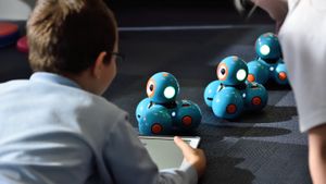 A boy lies on gray carpet operating a tablet, with several STEM education robots lined up in front of him and another person supervising