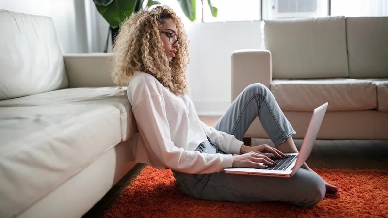 A woman sitting on the floor of a residence using her MacBook, with her back against a couch
