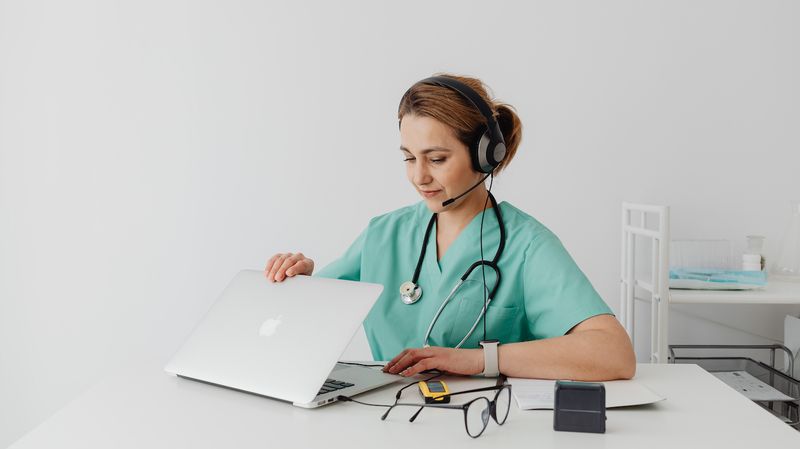 A woman wearing scrubs and a stethoscope sitting at a table and opening a Macbook