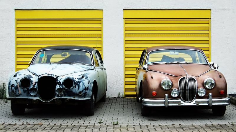 Two vintage cars sit side by side in front of bright yellow garage doors; the car on the left is in need of restoration while that on the right is in pristine condition