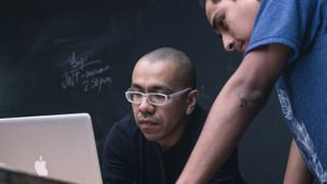 A man looking at a MacBook in front of a blackboard while another man looks over his shoulder