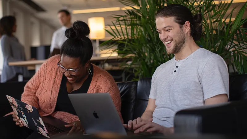 Smiling Jamf employees sit next to each other on a couch, working on MacBooks managed by Jamf.