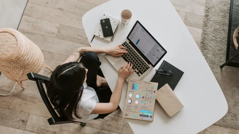 A worker with dark hair and a white T-shirt sits at a wooden desk. She is using a MacBook and iPhone and is photographed from above. She could be working from home or in a modern office. Jamf prepares you for whatever comes next in workplace management.