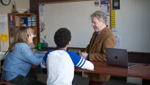 A teacher uses a MacBook provisioned by Installomator to aid two students in a classroom.