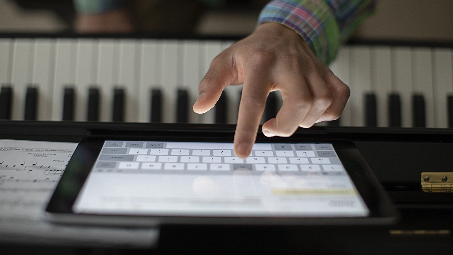 A child's hand types on an iPad that is propped up in front of a musical keyboard and sheet music.
