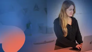 An employee with long light hair and complexion, wearking a black sweater, works on a MacBook managed by Jamf through declarative device management. The background is abstract colors.