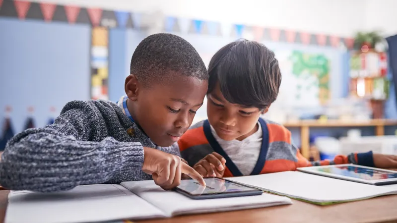 Two young students sit at a desk in a bright classroom, both leaning over the same iPad.