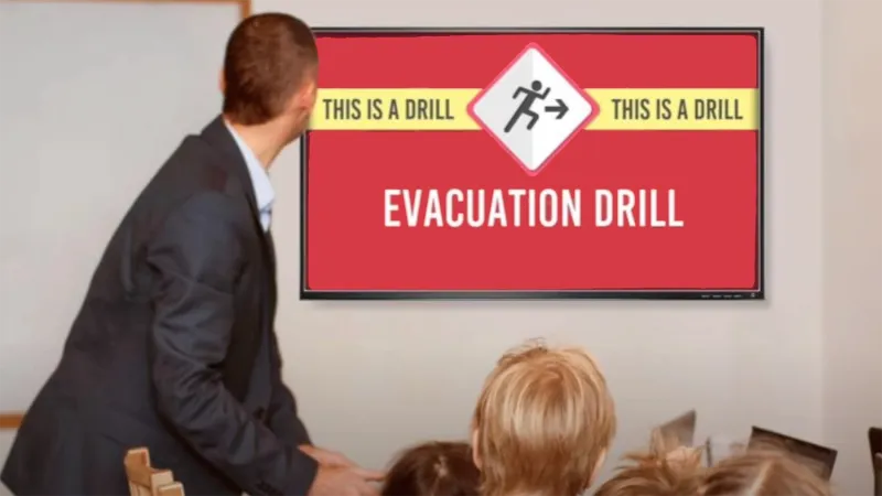 A teacher in a dark suit stands in a classroom with the tops of students' heads visible. They all look at signage which reads:
