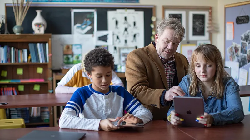 In a sunny classroom, a teacher helps two students on iPads that are managed by Jamf Pro and Apple Classroom.