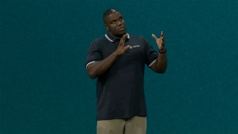 Alvin Bridges speaks animatedly, his hands making a shape in the air. He stand against a deep teal JNUC 2025 background. His black hair is cropped closely, and he wears a navy polo with khakis.