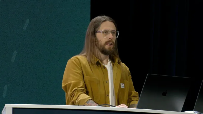 Anthony Darlow stands behind a podium and in front of a dark JNUC 2025 backdrop. He has long brown hair, a neatly-trimmed beard, and wears glasses. He wears a dark gold corduroy shirt pushed up at the sleeves over a white T-shirt.