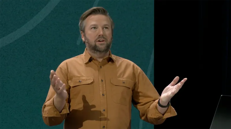 Eric Blakely stands in front of a JNUC 2025 backgrounds. He has swept-back grown hair, a brown beard, and gestures with both hands. He is wearing a dark gold corduroy shirt pushed up at the sleeves.