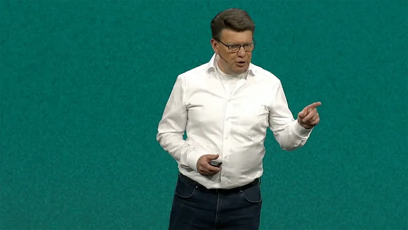 Ernout van Beek stands in front of a teal JNUC 2025 background, gesturing as he speaks. He wears a white dress shirt and dark pants. He has a wave of brown hair and wears brown glasses.